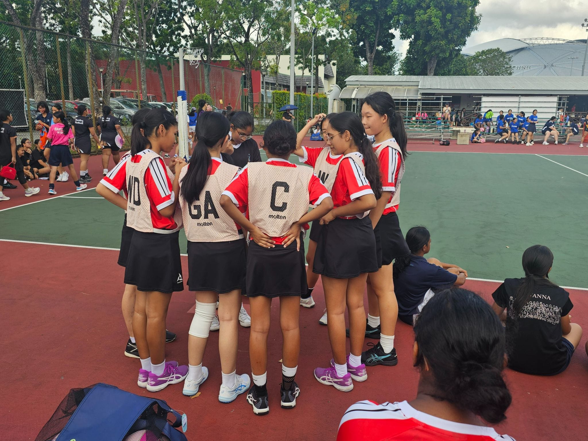 Young girls in red and white netball bibs huddle together on an outdoor court during a tournament.