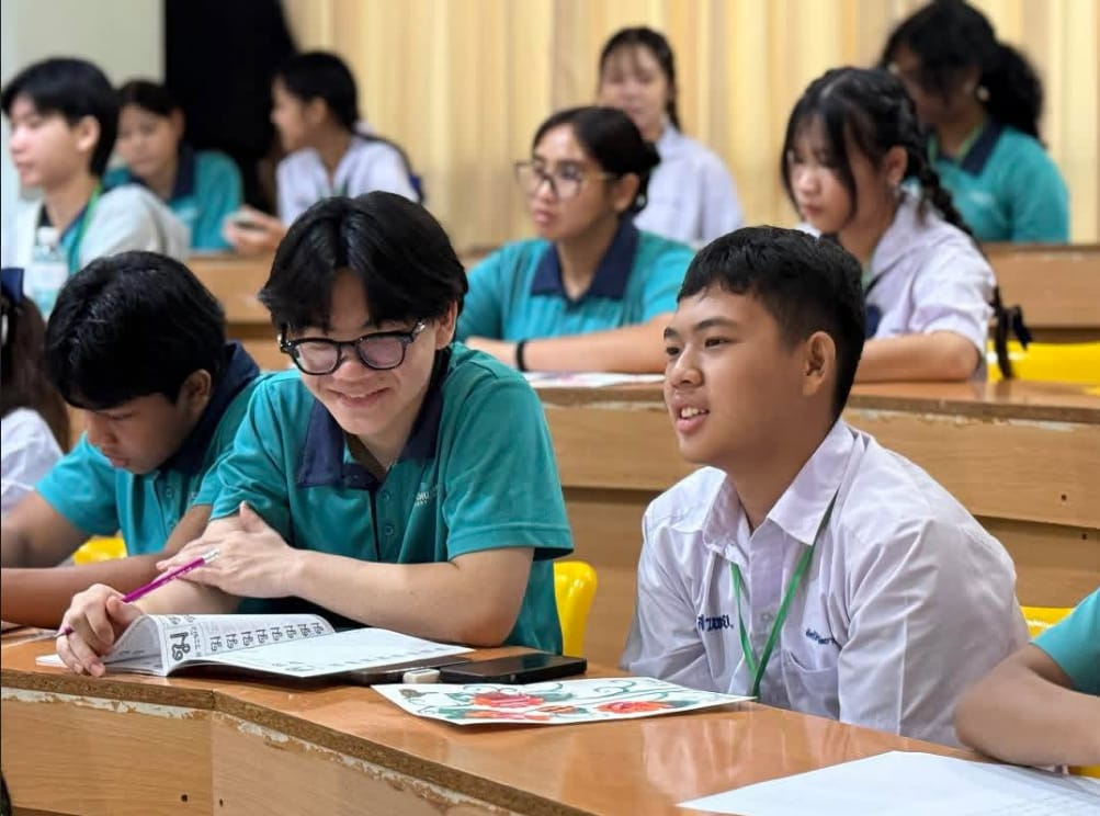 Students in teal uniforms sit at classroom desks, with two boys in the foreground smiling and looking forward.