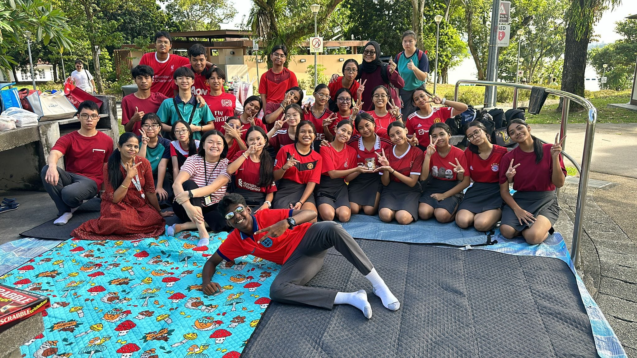Large group of students mostly in red jerseys posing outdoors on picnic mats, making peace signs, with trees behind them.