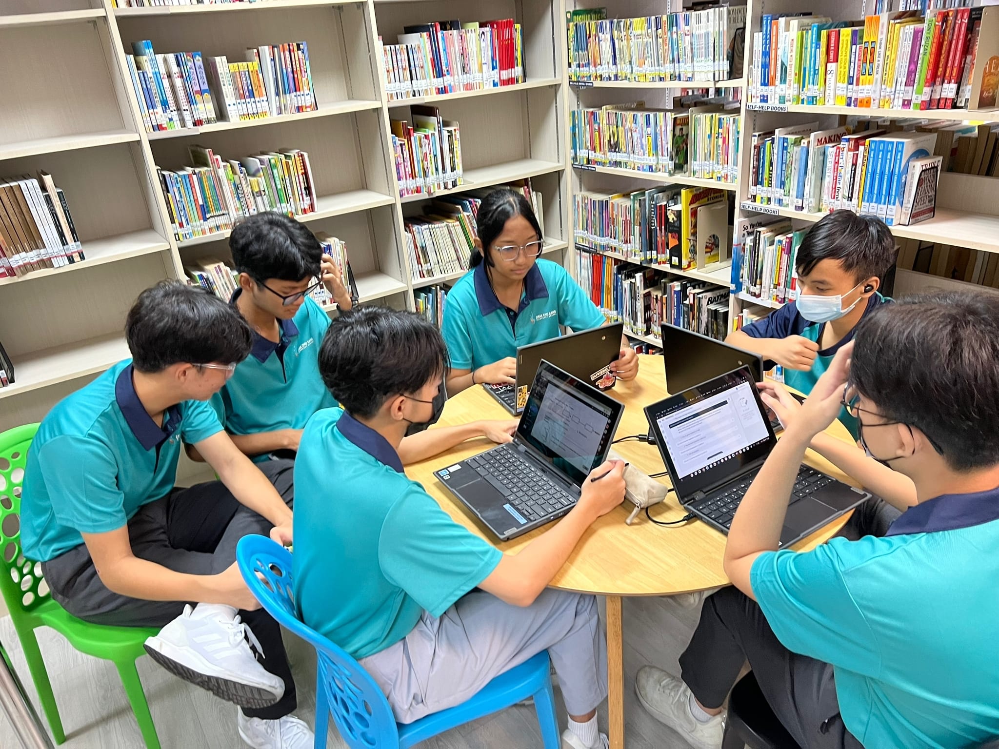 Six students in teal uniforms sit around a table with laptops in a library filled with bookshelves.