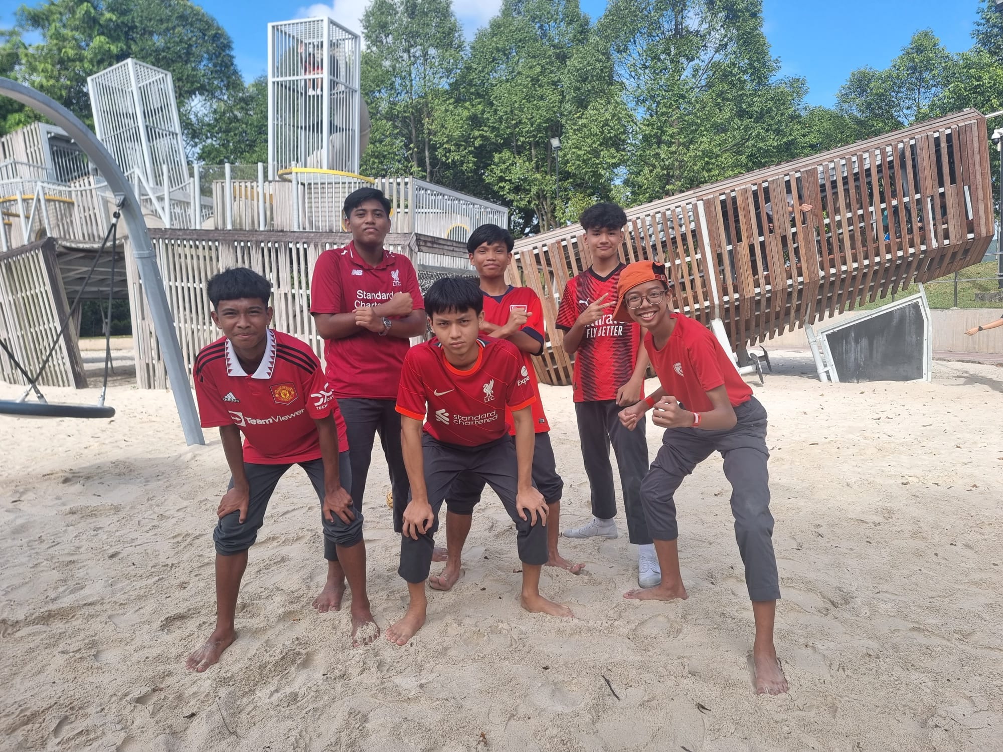 Six teenage boys in red football jerseys posing barefoot on sand at an outdoor playground with wooden structures.