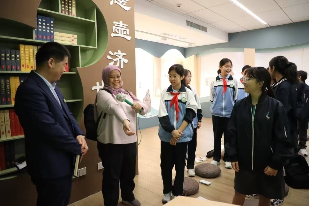 A woman in a hijab holds a microphone toward students in school uniforms inside a room with Chinese text and bookshelves.