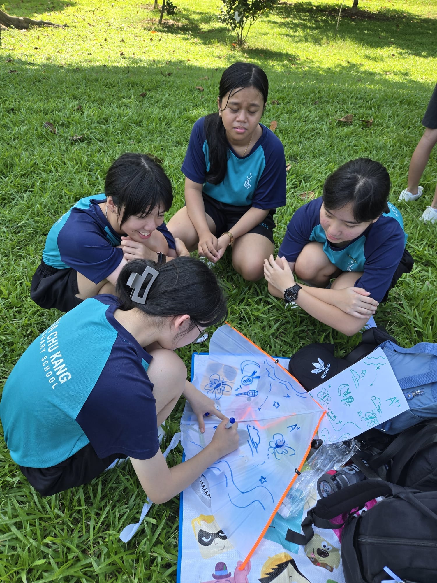 Four students in school uniforms sit on grass, drawing colorful designs on a kite outdoors.
