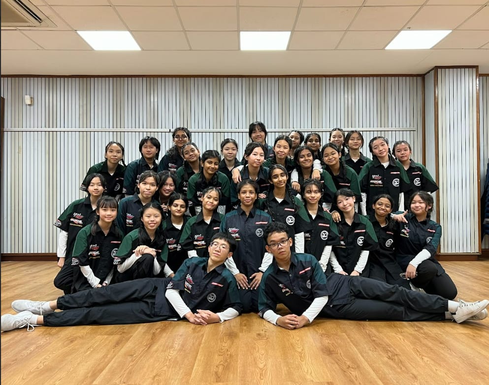 A large group of young students in matching dark green and black uniforms pose together in a indoor room.