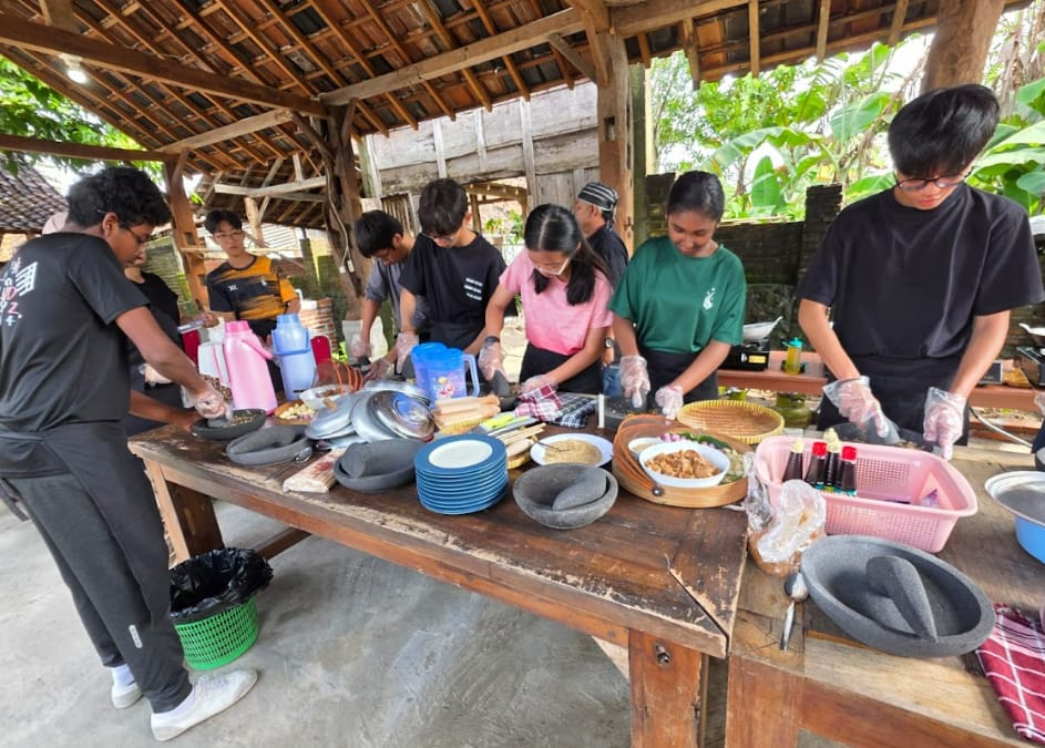 Several young people wearing gloves prepare food at a large wooden table under an open-air thatched shelter.