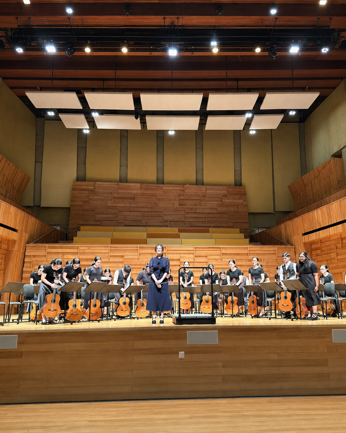 A conductor in blue stands center stage with a large youth guitar ensemble on a wood-paneled concert hall stage.