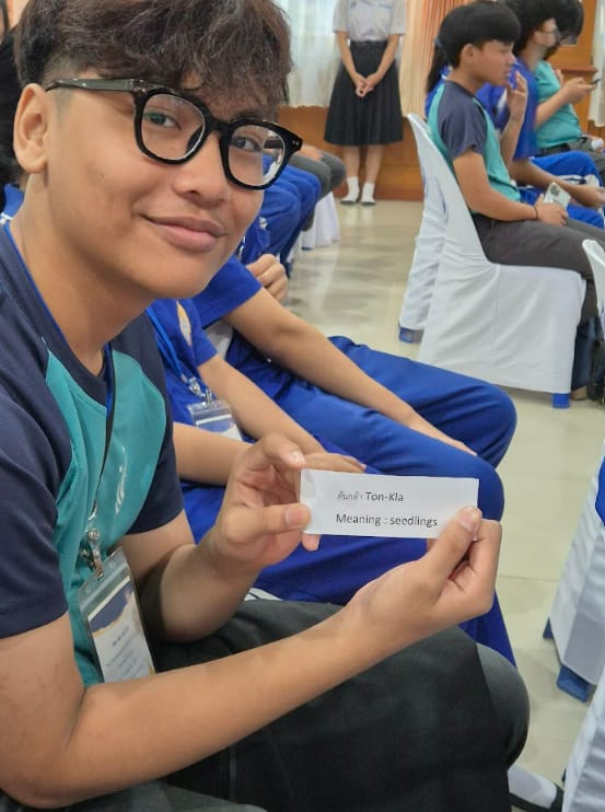 Young person with glasses holding a card reading "Ton-Kla, Meaning: seedlings" in a room with others.