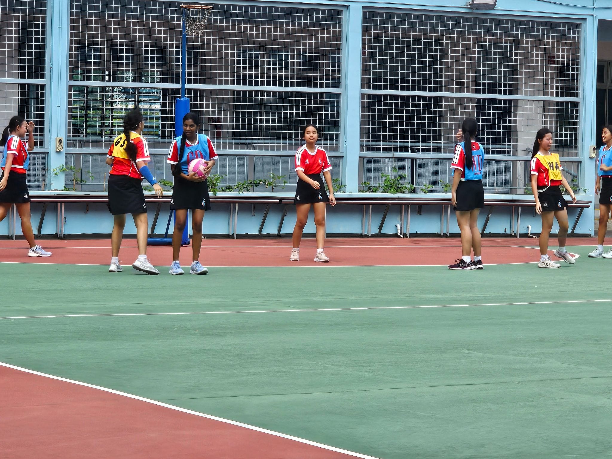 Girls in red uniforms play netball on an outdoor court, one holding a pink ball near a blue goal post.