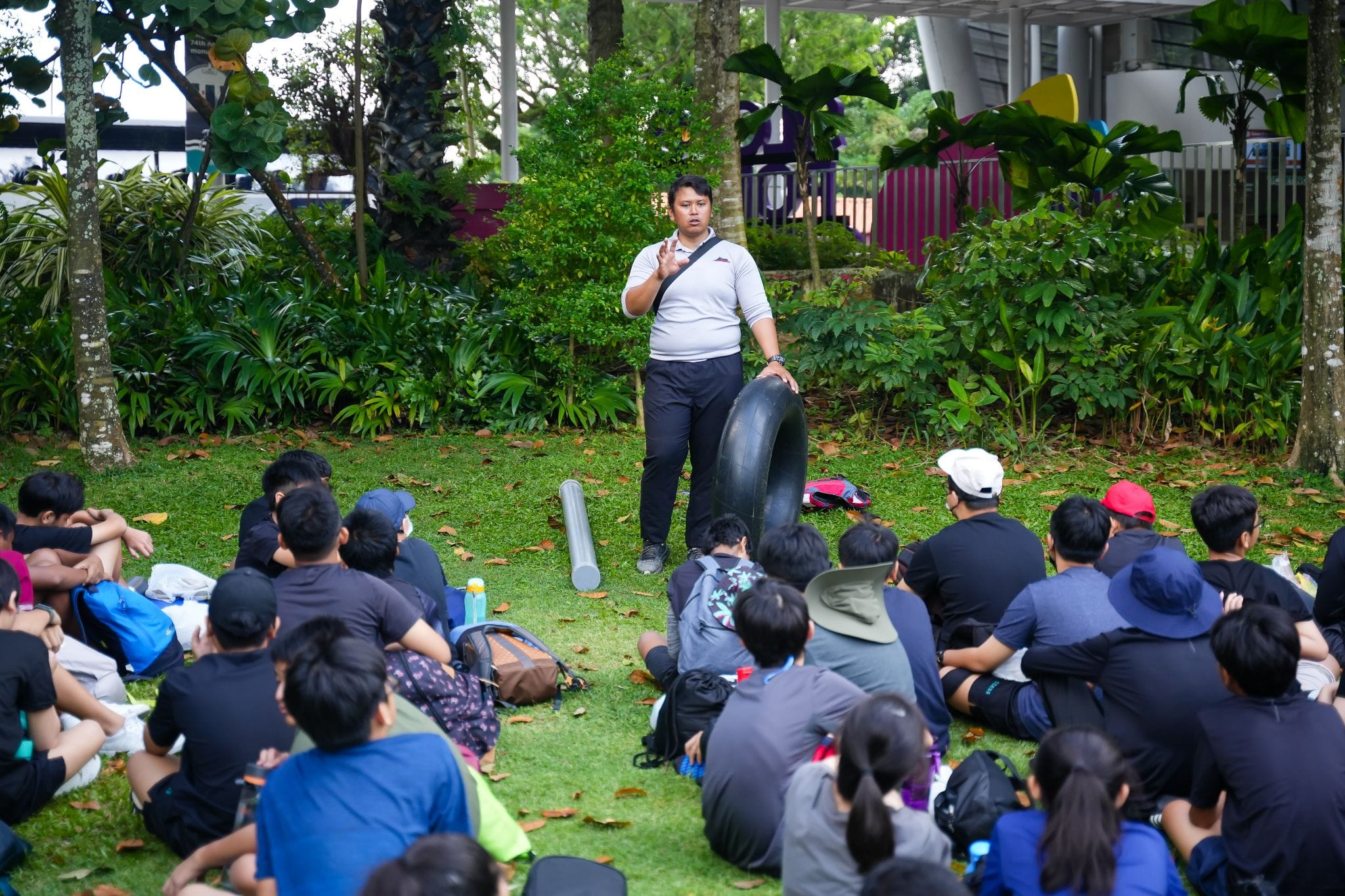A man holding an inflatable tube speaks to a large group of students sitting on grass outdoors near trees.