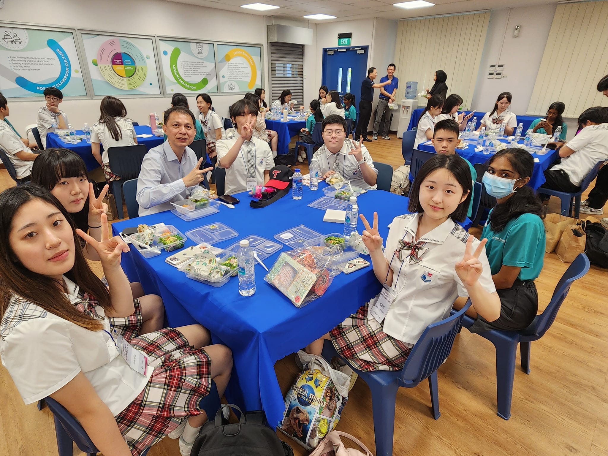 Students in school uniforms sit at blue-clothed tables with food, posing with peace signs in a bright classroom.