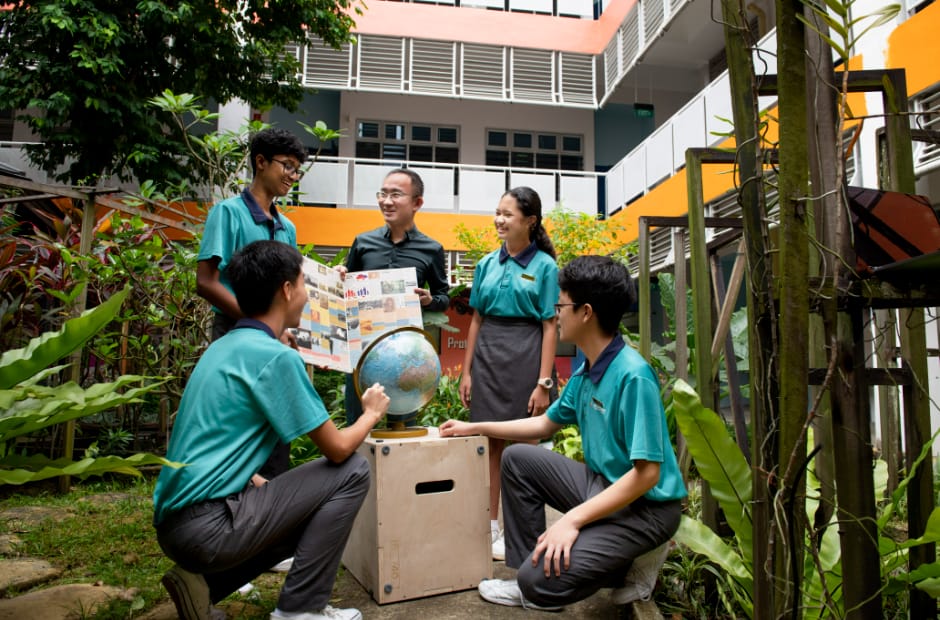 Five students in teal uniforms gather outdoors around a globe, with a teacher holding a poster near a school building.