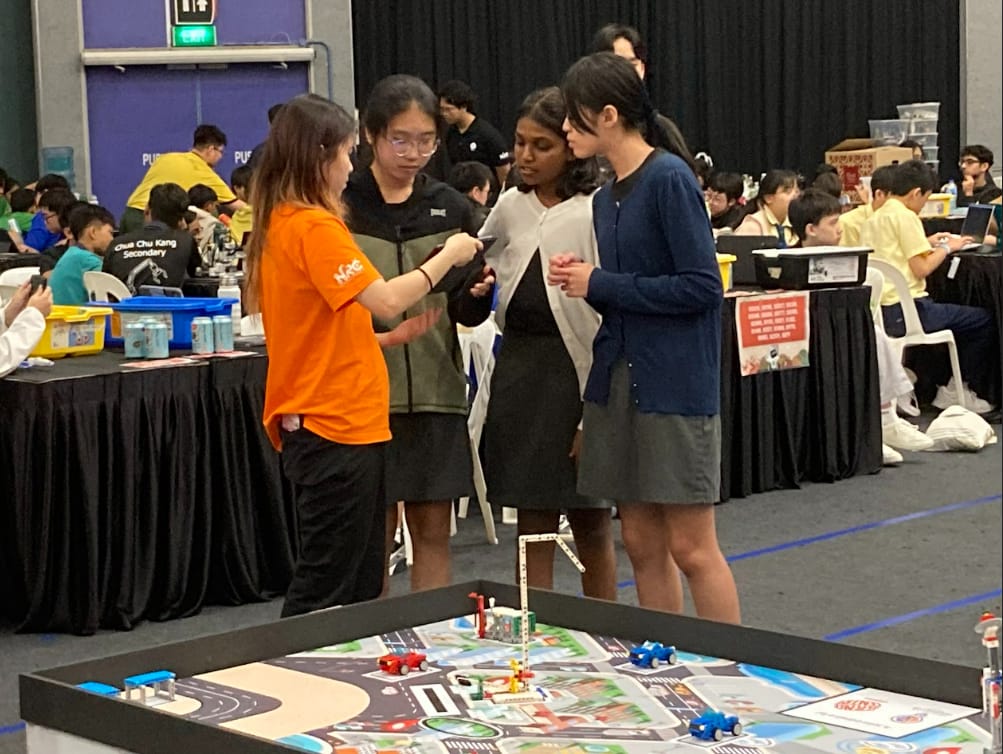 Four students gather around a robotics competition mat with toy cars and structures in a busy hall.