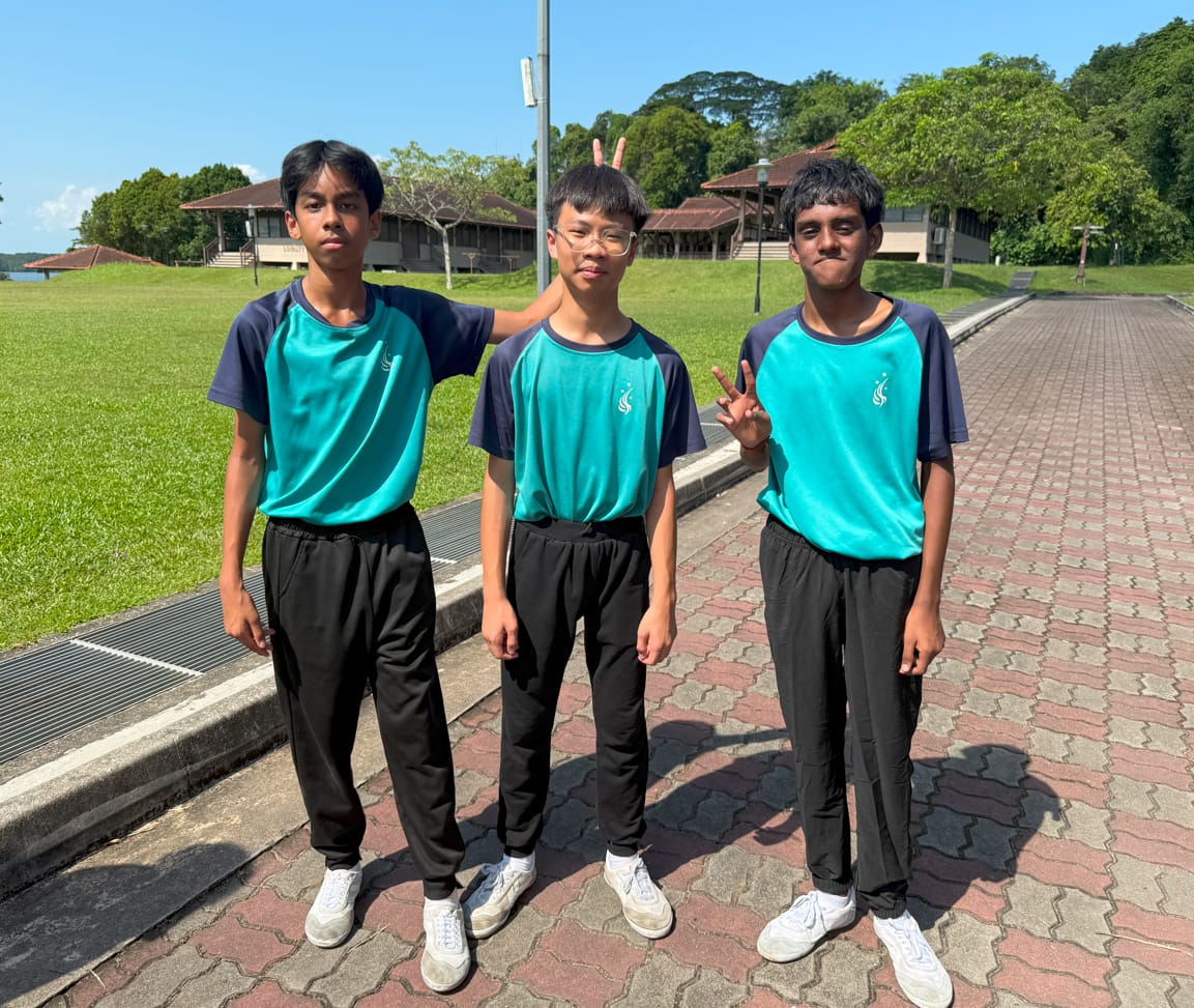 Three teenage boys in matching teal and navy uniforms stand on a brick path outdoors, one making a peace sign.