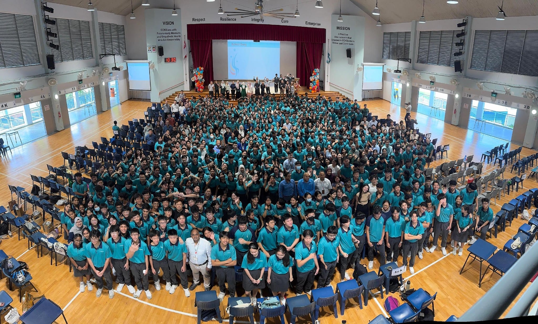 Large group of students in teal uniforms gathered in a school hall for a group photo, with a stage behind them.