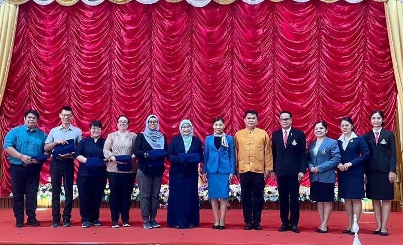 Ten people stand in a row on a red-carpeted stage before a large red curtain, some holding dark gift boxes.