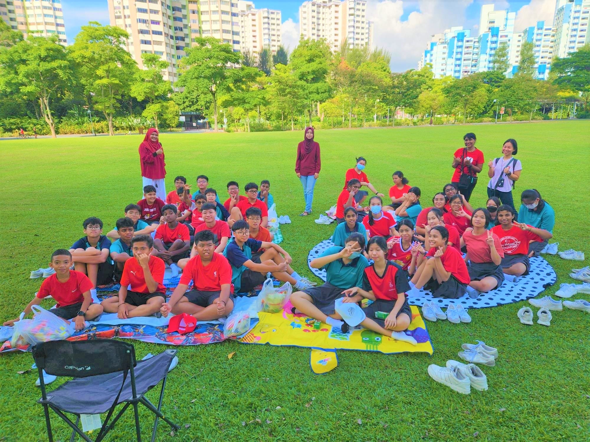 A large group of students in red shirts sit on picnic mats on a green field with tall apartment buildings behind them.