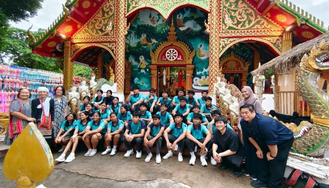 A large group of students in teal shirts poses in front of a colorful ornate Thai Buddhist temple.