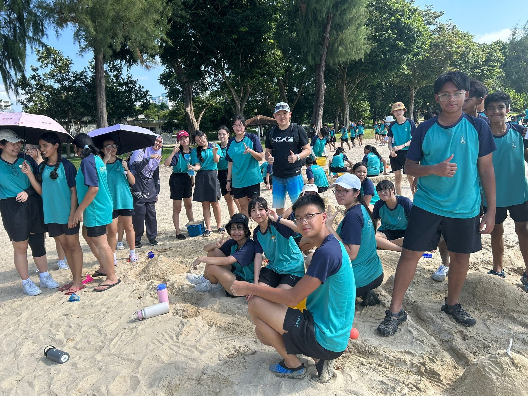 A large group of students in teal uniforms pose and smile on a sandy beach surrounded by trees.