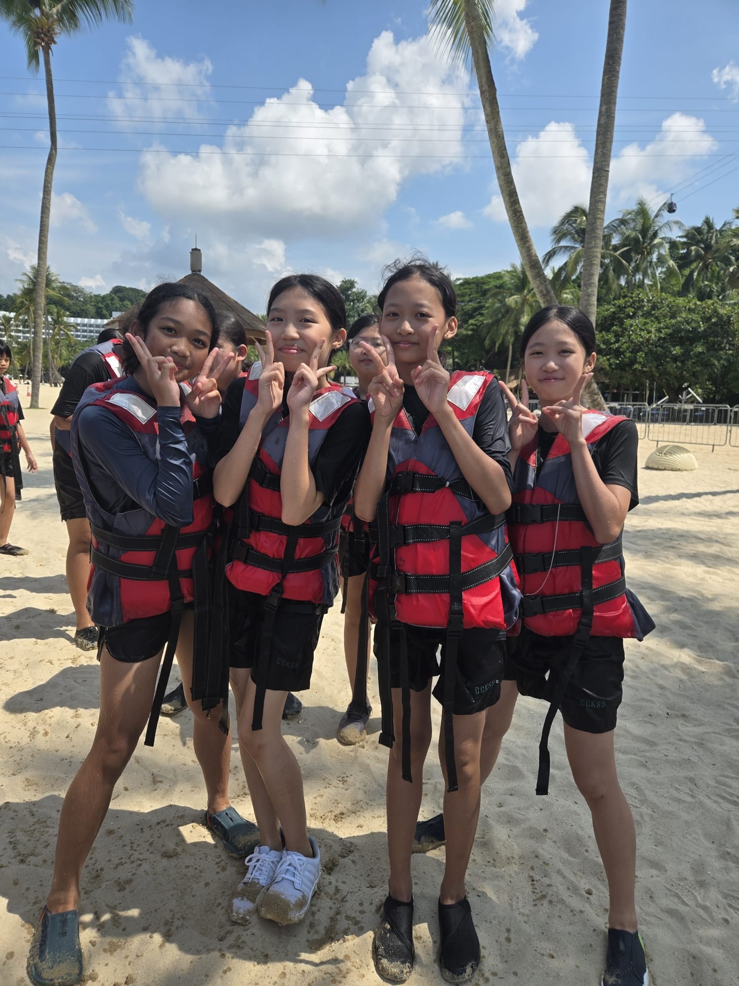 Four girls wearing red life jackets pose with peace signs on a sandy beach lined with palm trees.