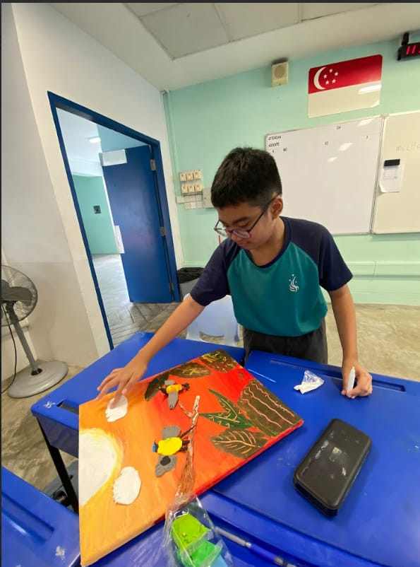A boy with glasses works on a colorful painting of birds and plants on a blue table in a classroom.