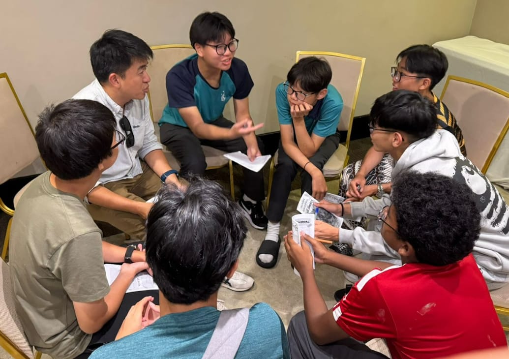 Seven young people sit in a circle on chairs and the floor, talking and holding papers in a room.