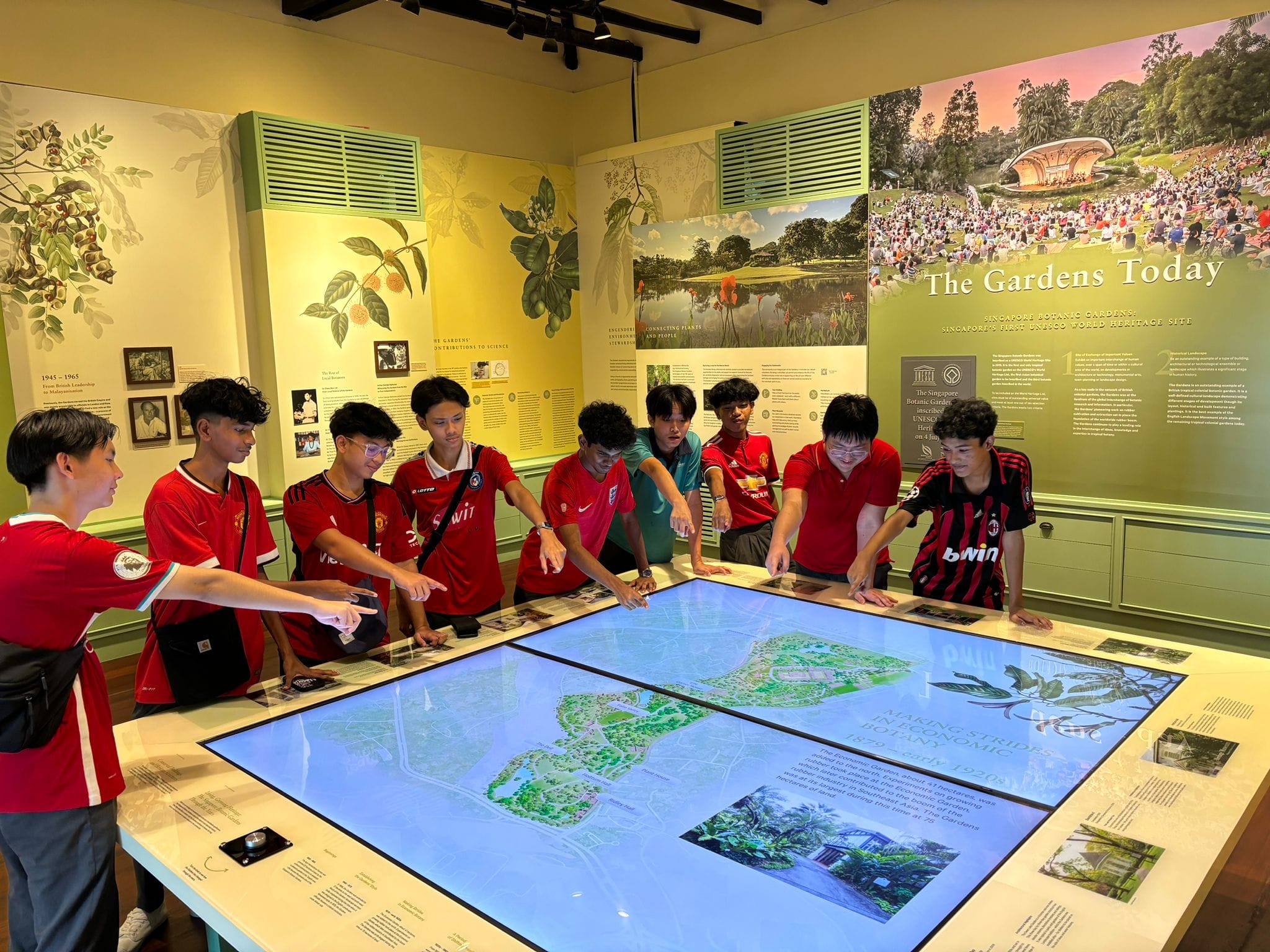 A group of teenagers in red jerseys gather around a large interactive map display inside a botanic gardens museum.
