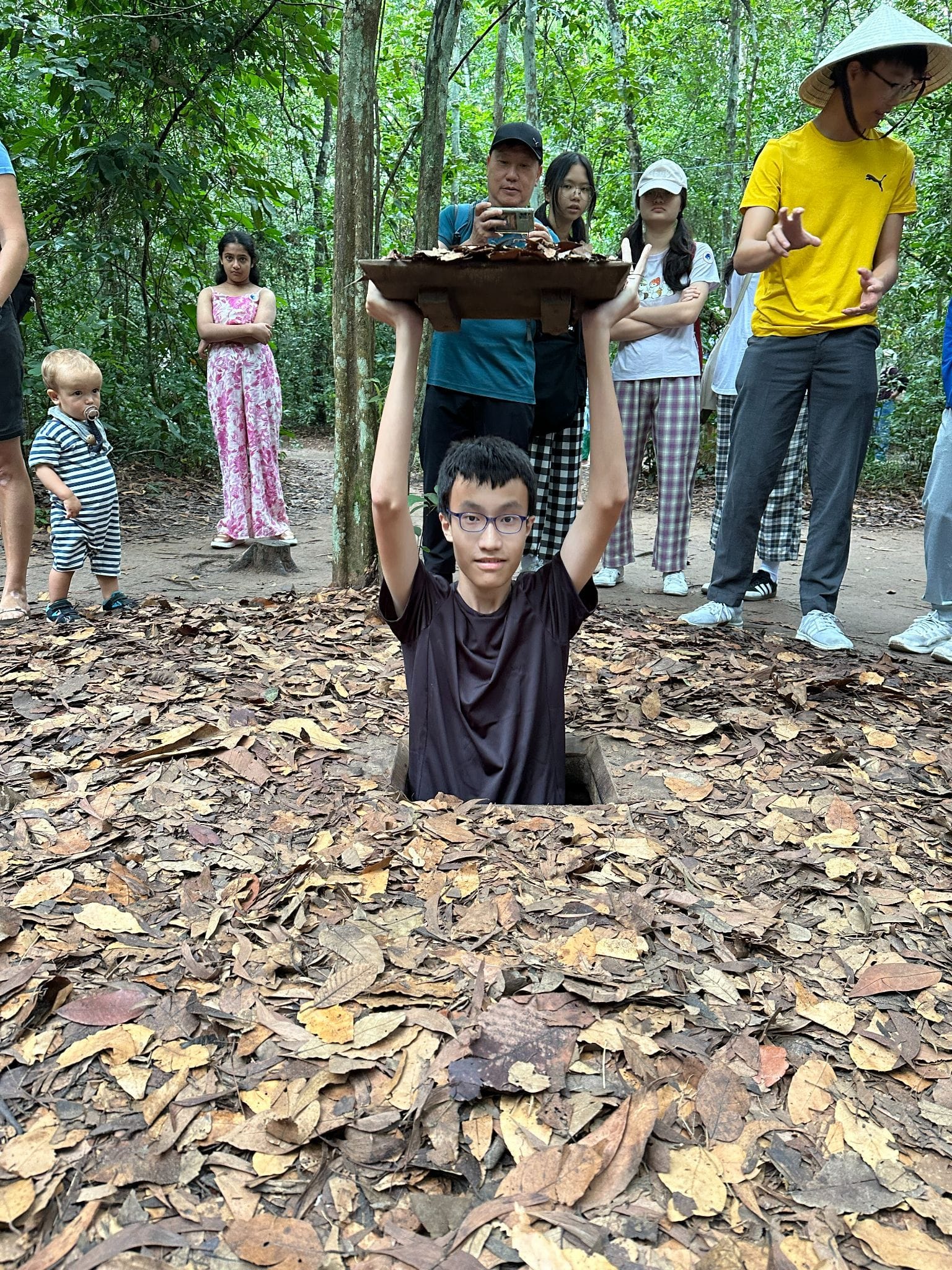 A teen with glasses emerges from a small underground tunnel entrance covered in dry leaves, lifting the camouflaged lid overhead.