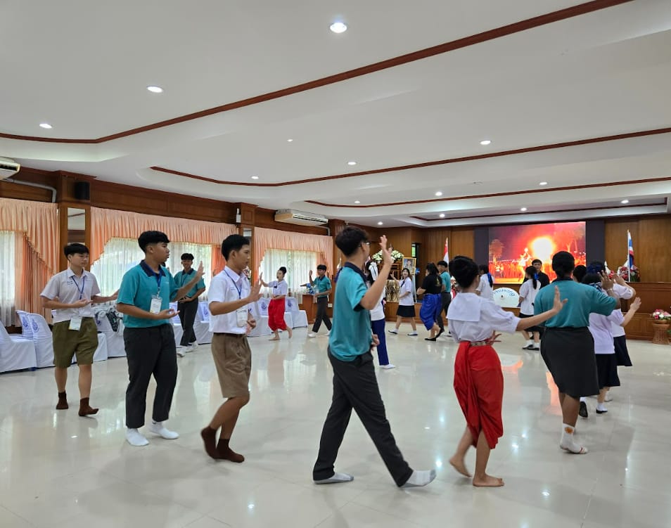 A group of young people in teal shirts and school uniforms practice traditional Thai dance in a large indoor hall.