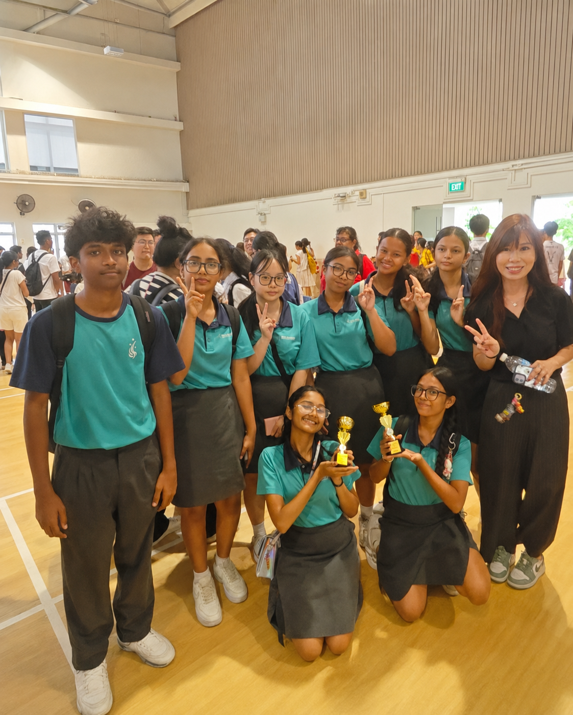 Nine students in teal uniforms and a teacher pose together, with two holding trophies.