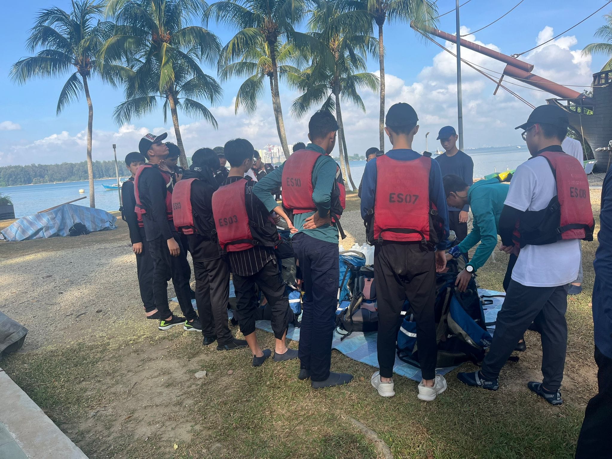 A group of young people wearing red life vests gather near a beach with palm trees and gear on the ground.