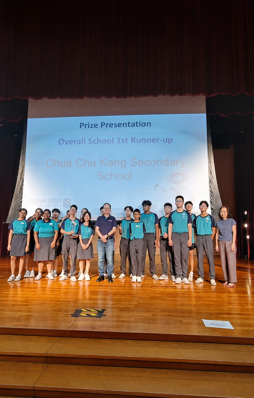 Students in teal uniforms pose on stage for a Prize Presentation, named Overall School 1st Runner-up for Chua Chu Kang Secondary School.
