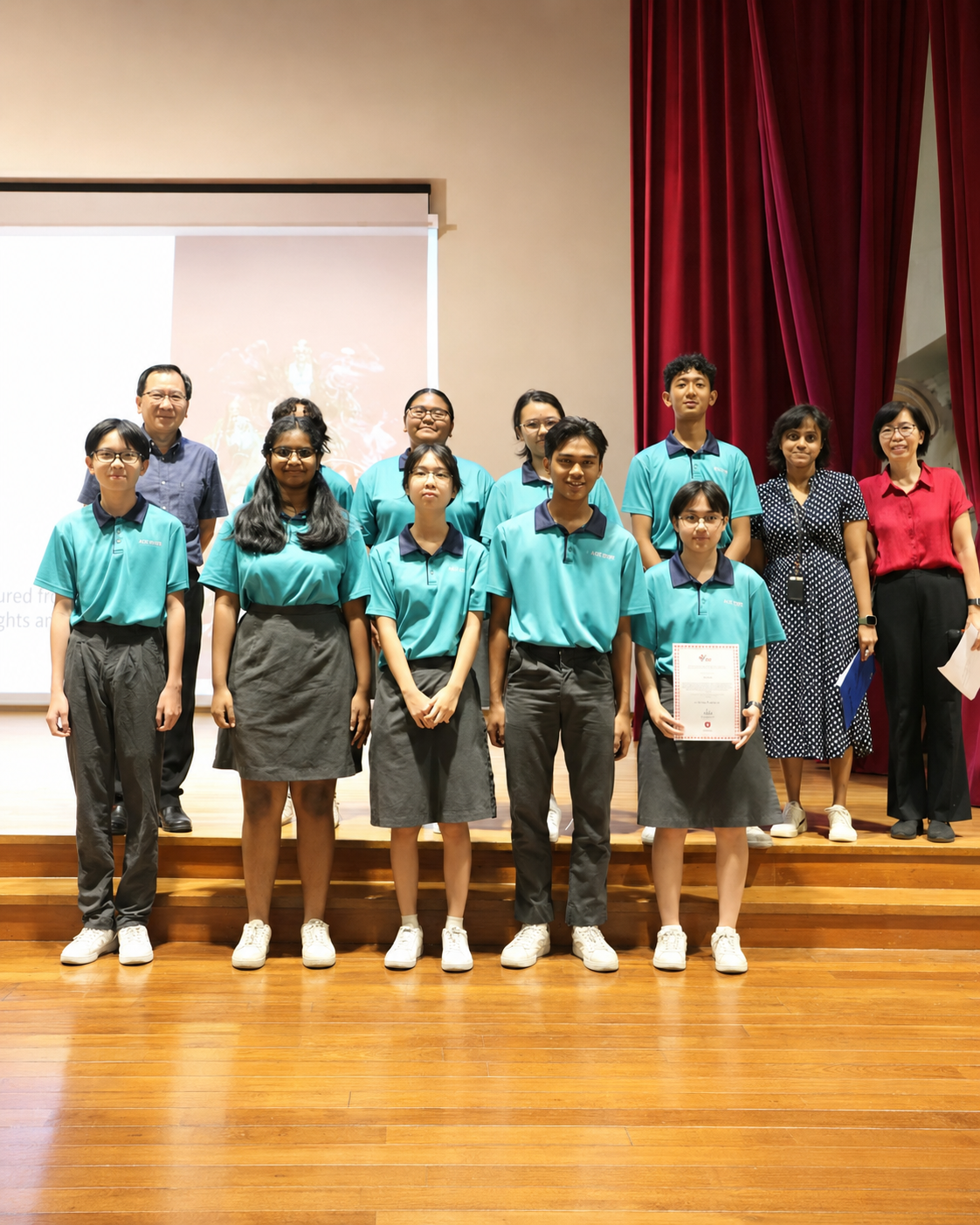 A group of students in teal uniforms pose with two adult teachers, one student holding a certificate.