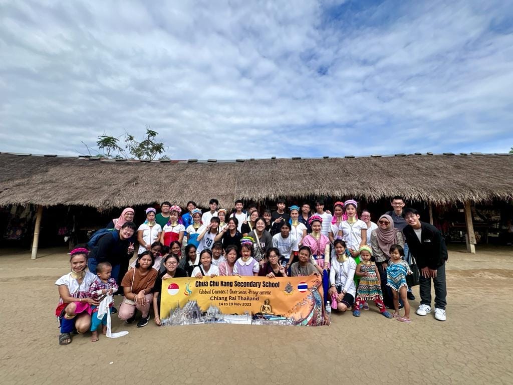 Large group poses with a Chua Chu Kang Secondary School Chiang Rai Thailand 2023 banner in front of a thatched building.