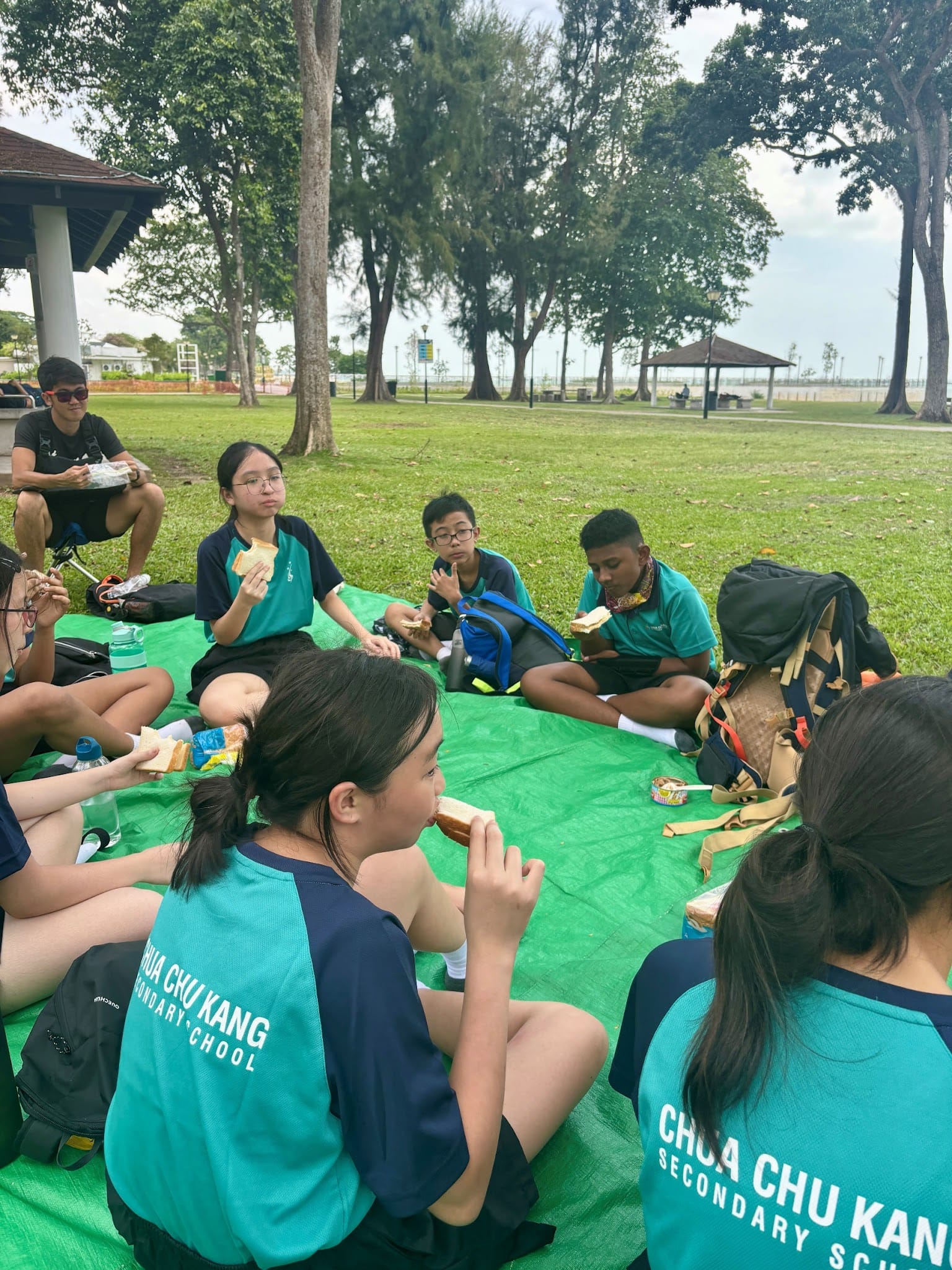 Students in Chua Chu Kang Secondary School uniforms sit in a circle on a green mat in a park, eating bread.