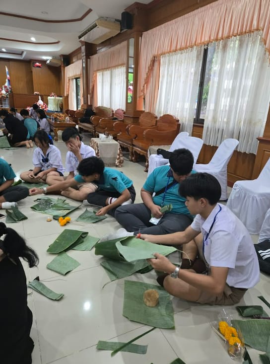 Students in uniforms sit on a tiled floor working with large green banana leaves and flowers indoors.