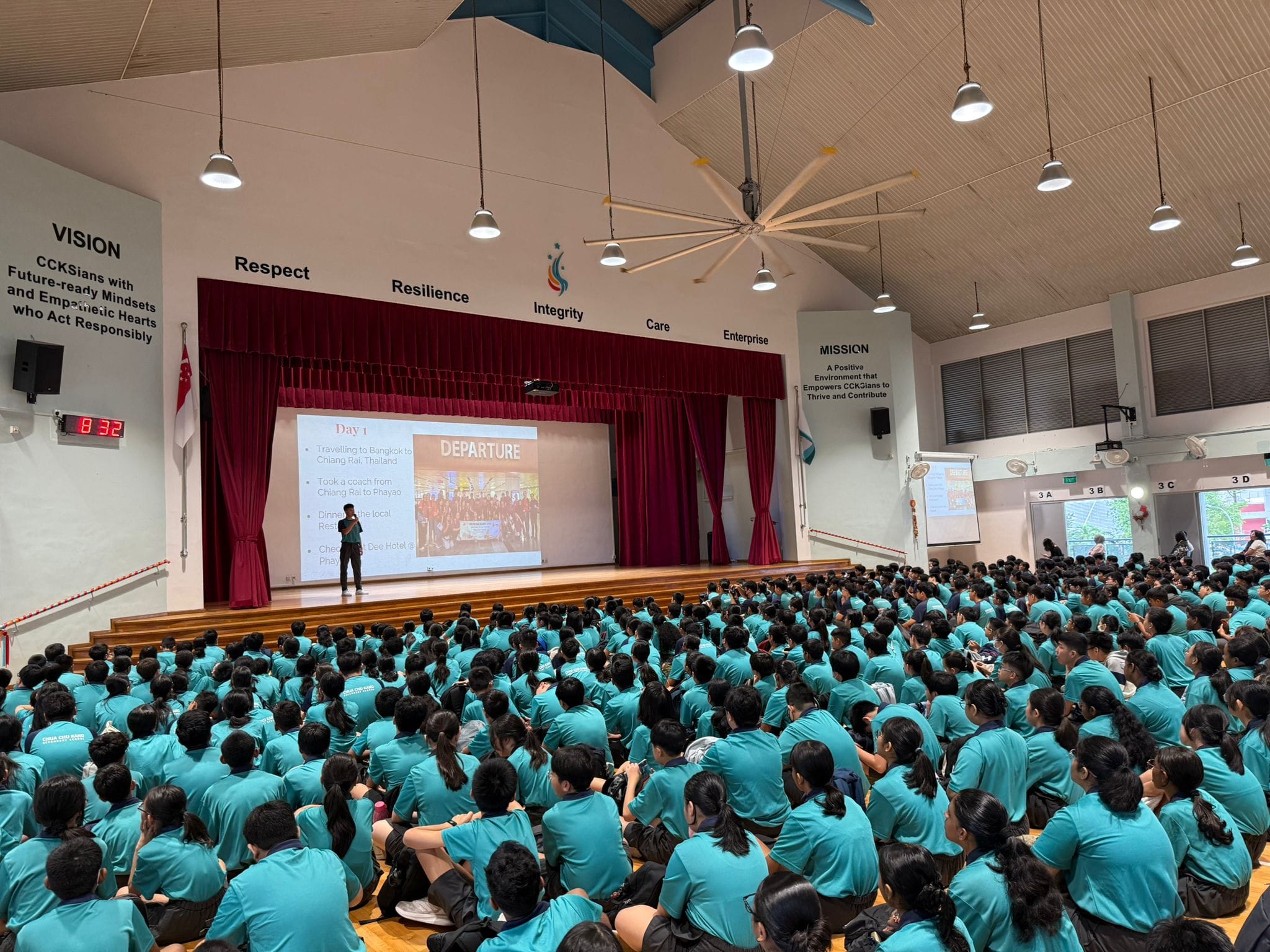 Students in teal uniforms sit on a school hall floor facing a stage with a Departure slideshow presentation.