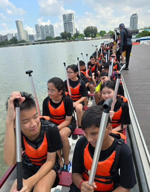 A group of young people in orange life vests sit in a long dragon boat on a river, holding paddles.