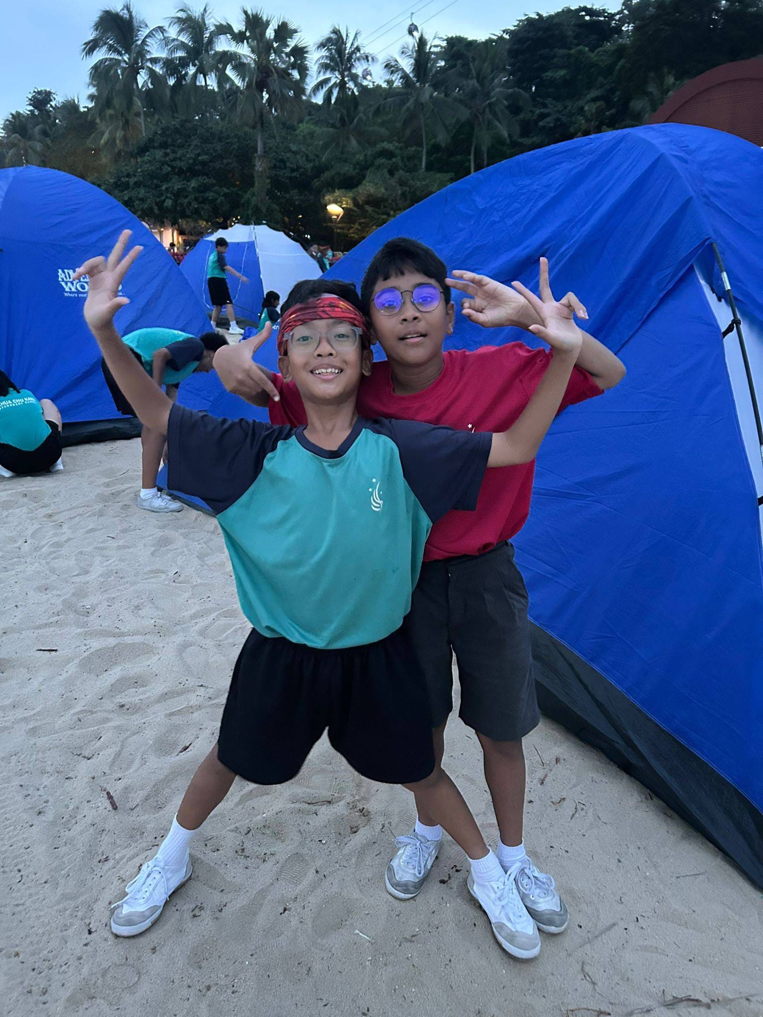 Two boys in teal and red uniforms pose together smiling near blue camping tents in an indoor venue.