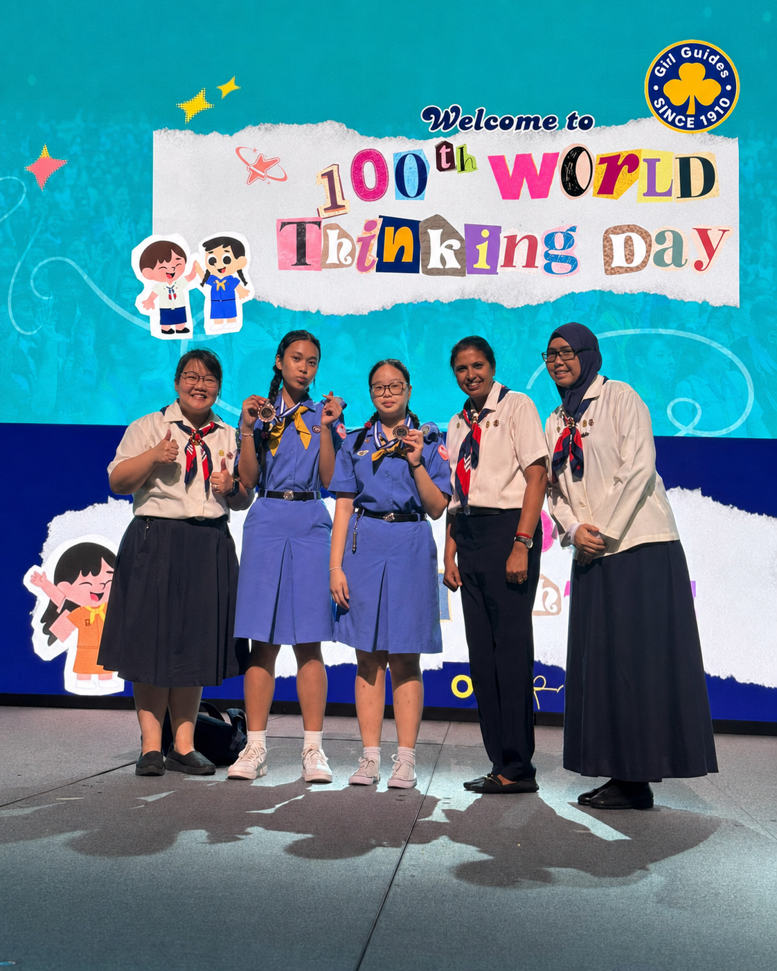 Five girls in scout uniforms pose on stage, two holding medals, before a "Welcome to 100th World Thinking Day" banner.