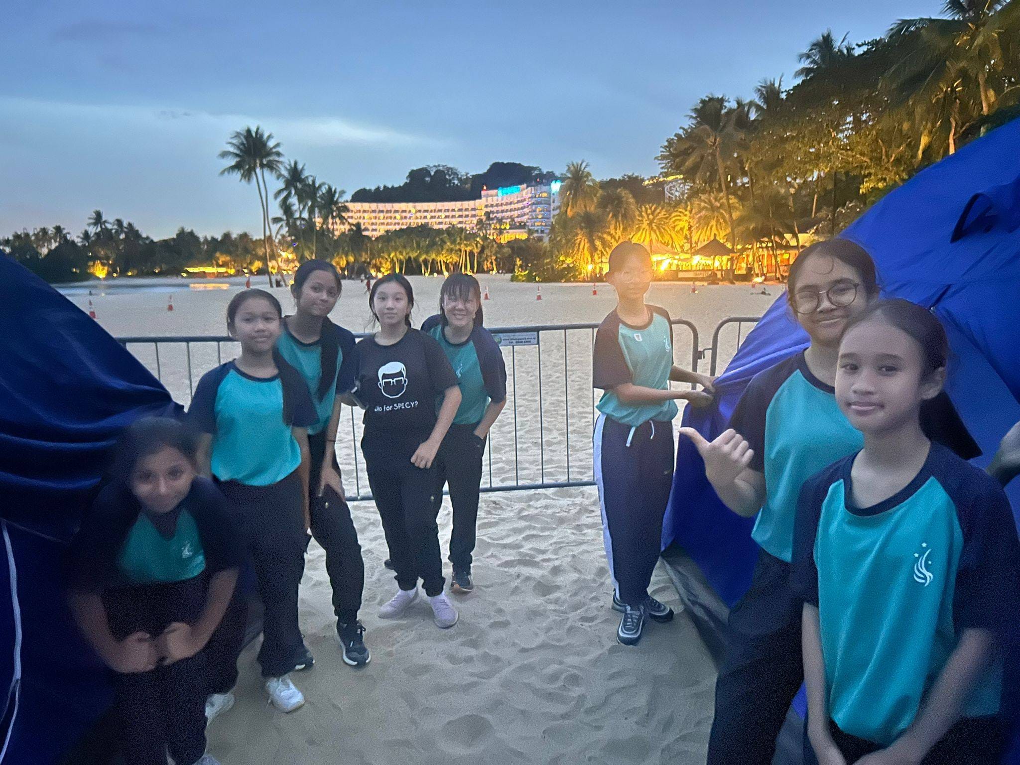 Eight girls in matching teal uniforms pose together smiling near blue camping tents in an indoor venue.
