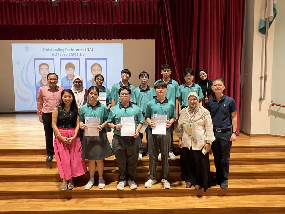 Group of students in teal uniforms holding certificates on a stage, with teachers, beside a slideshow reading Outstanding Performers.