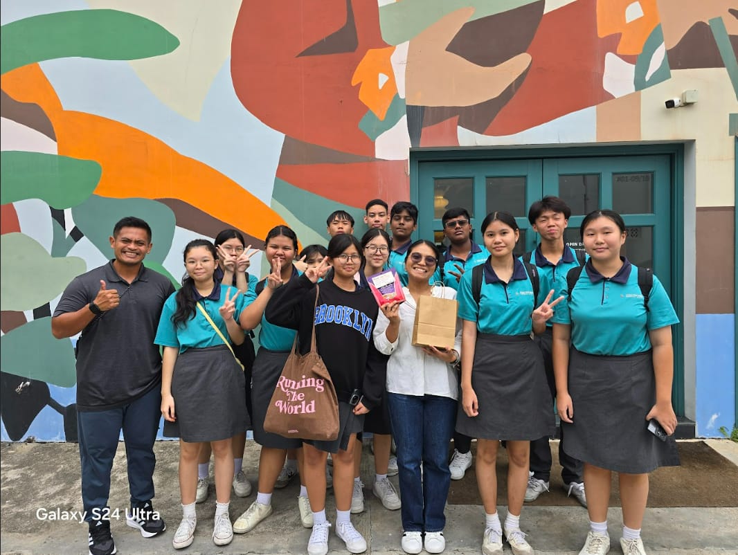 A group of students in teal uniforms and two adults pose smiling in front of a colorful painted mural wall.