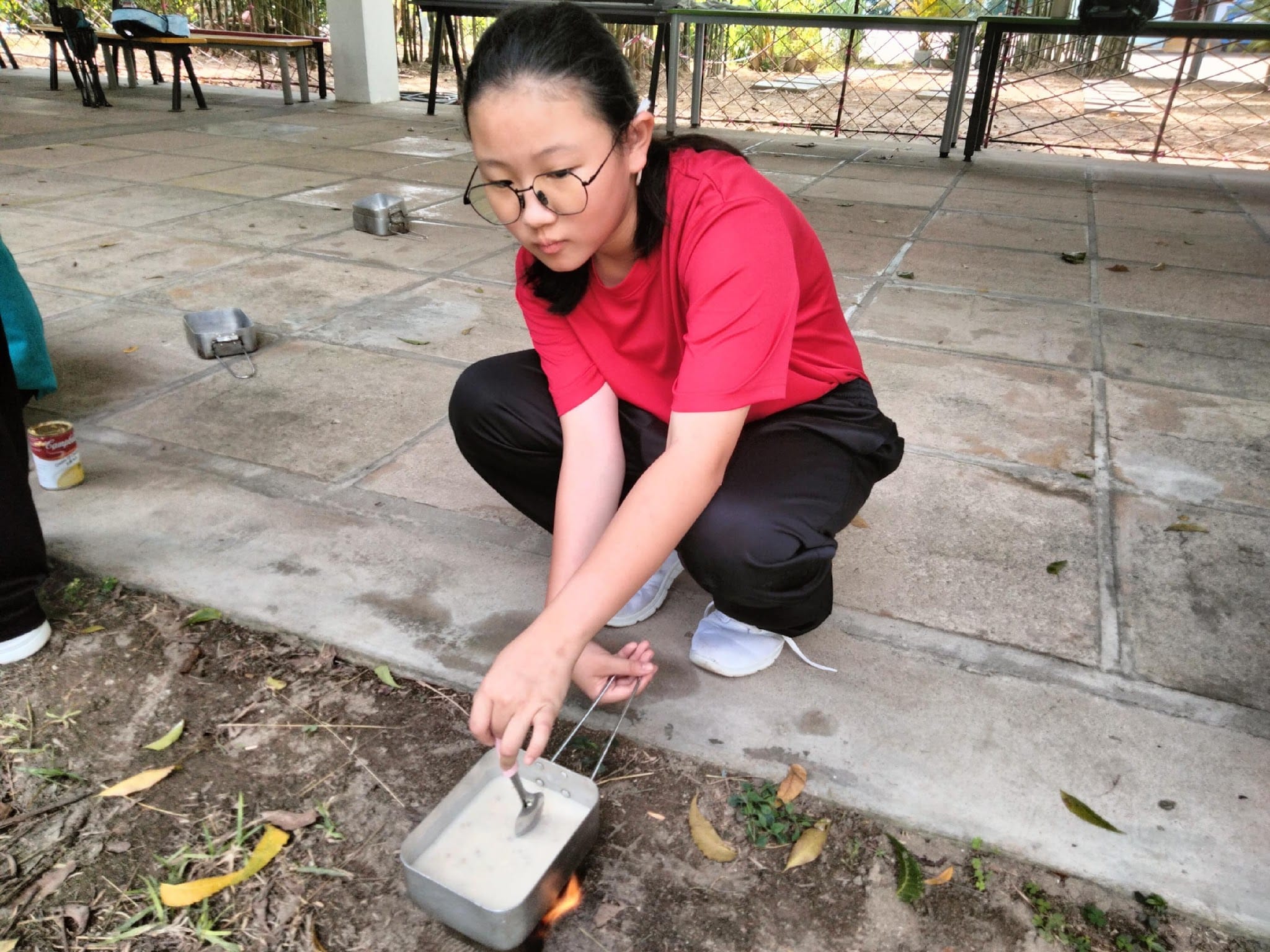 A young girl in a red shirt and glasses crouches outdoors, heating a small metal pot over an open flame.
