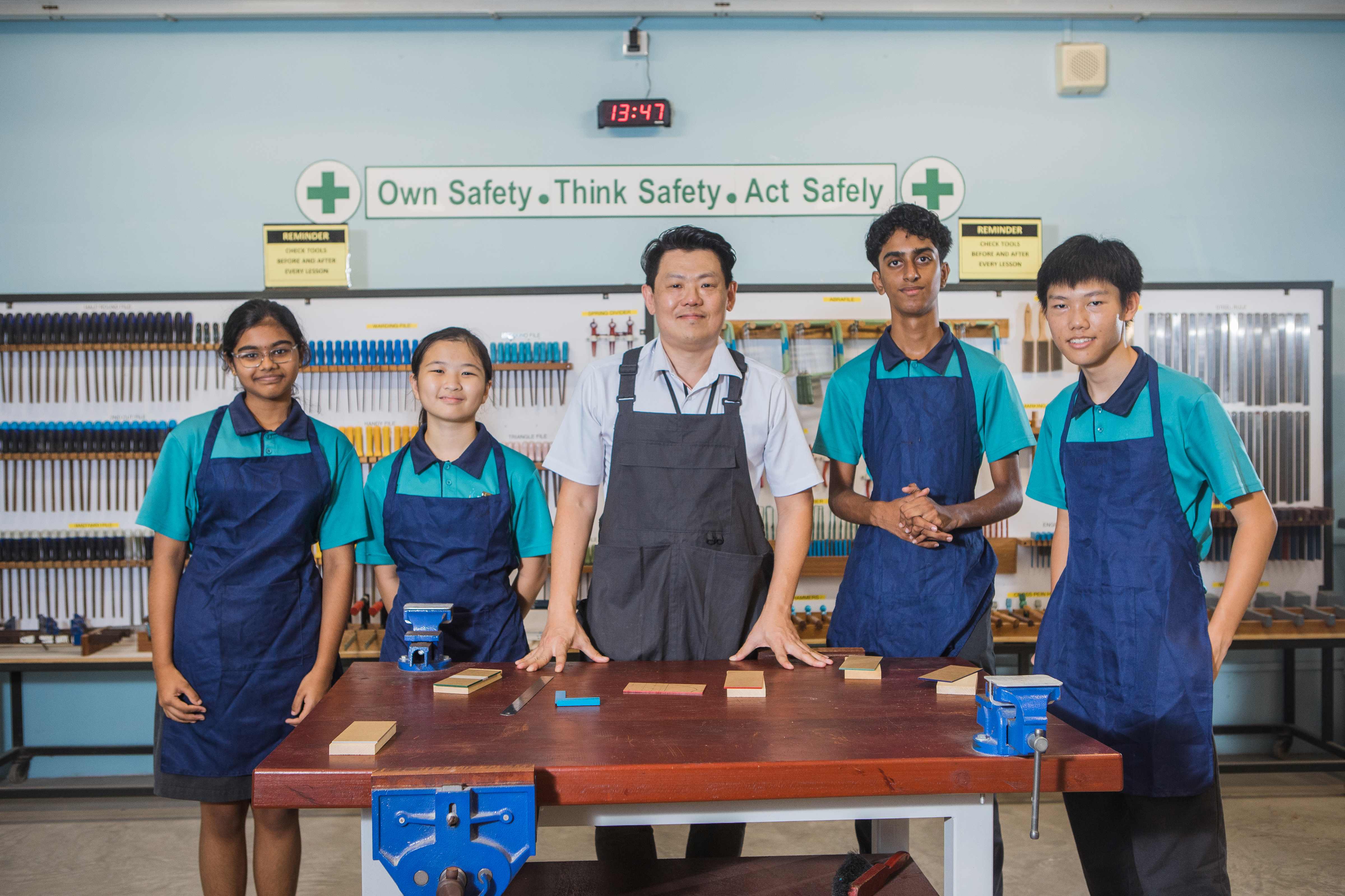 A teacher and four students in aprons stand at a workshop bench in a school workshop with tools on the wall.