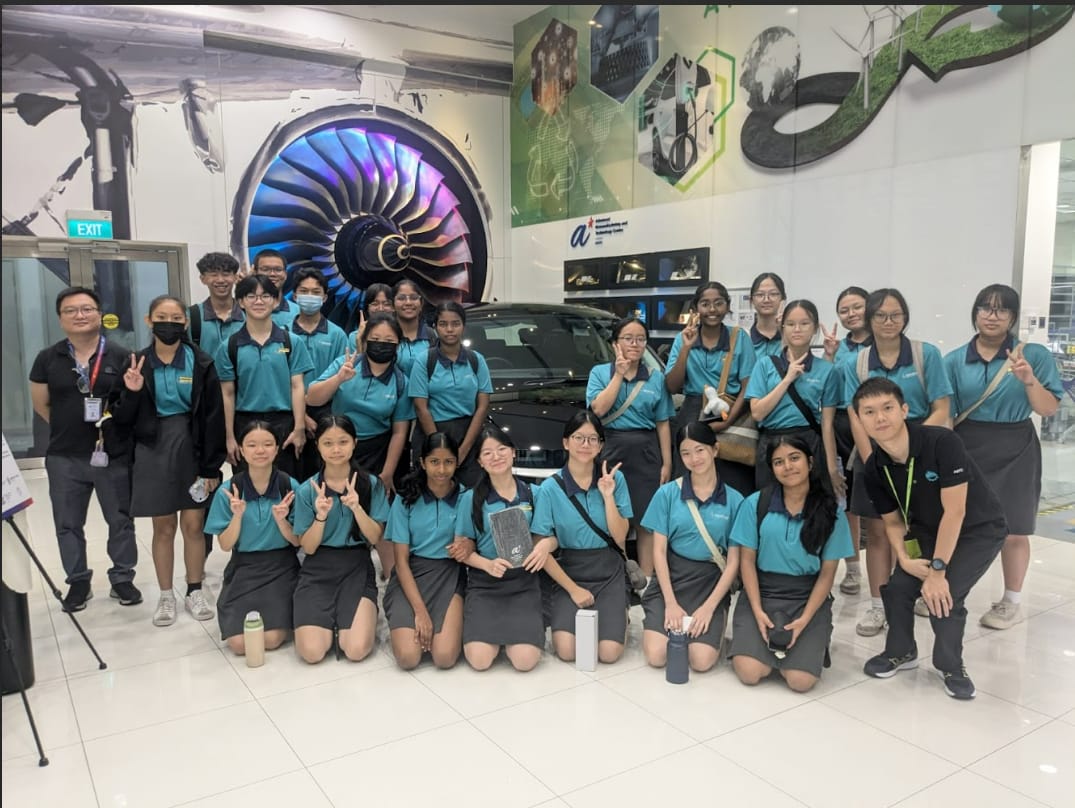 A group of students in teal polo shirts poses in front of a jet engine display and a dark car indoors.