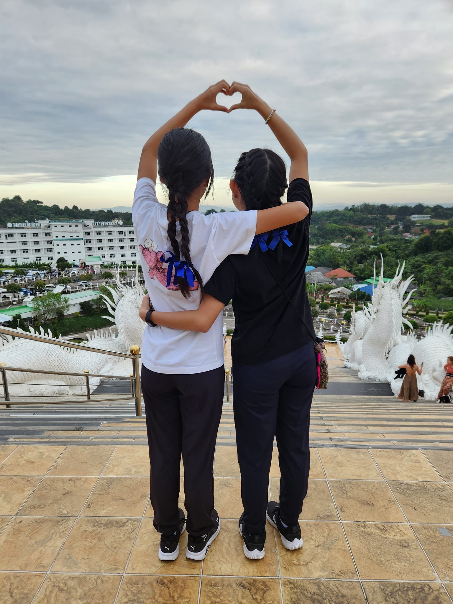 two girls make a heart-shape while looking over the distant horizon.