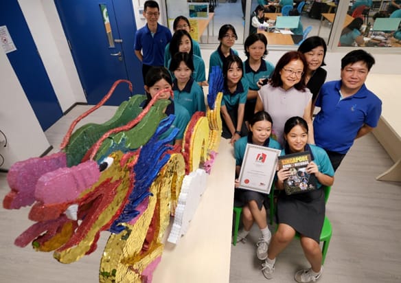 A group of students and teachers pose with a colorful dragon sculpture and two students hold a certificate and book.