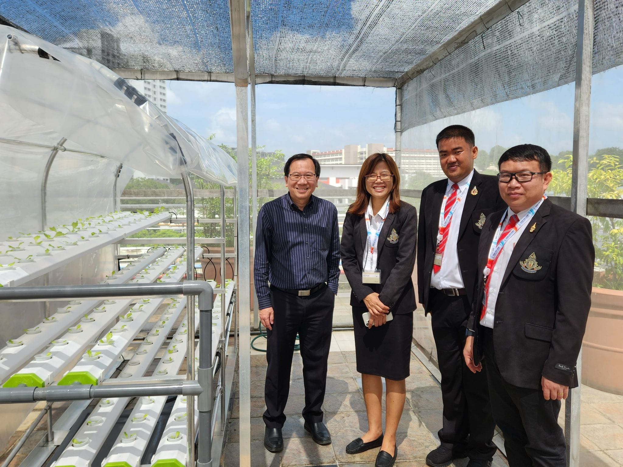 Four people in formal attire stand smiling beside hydroponic growing trays inside an outdoor rooftop greenhouse structure.