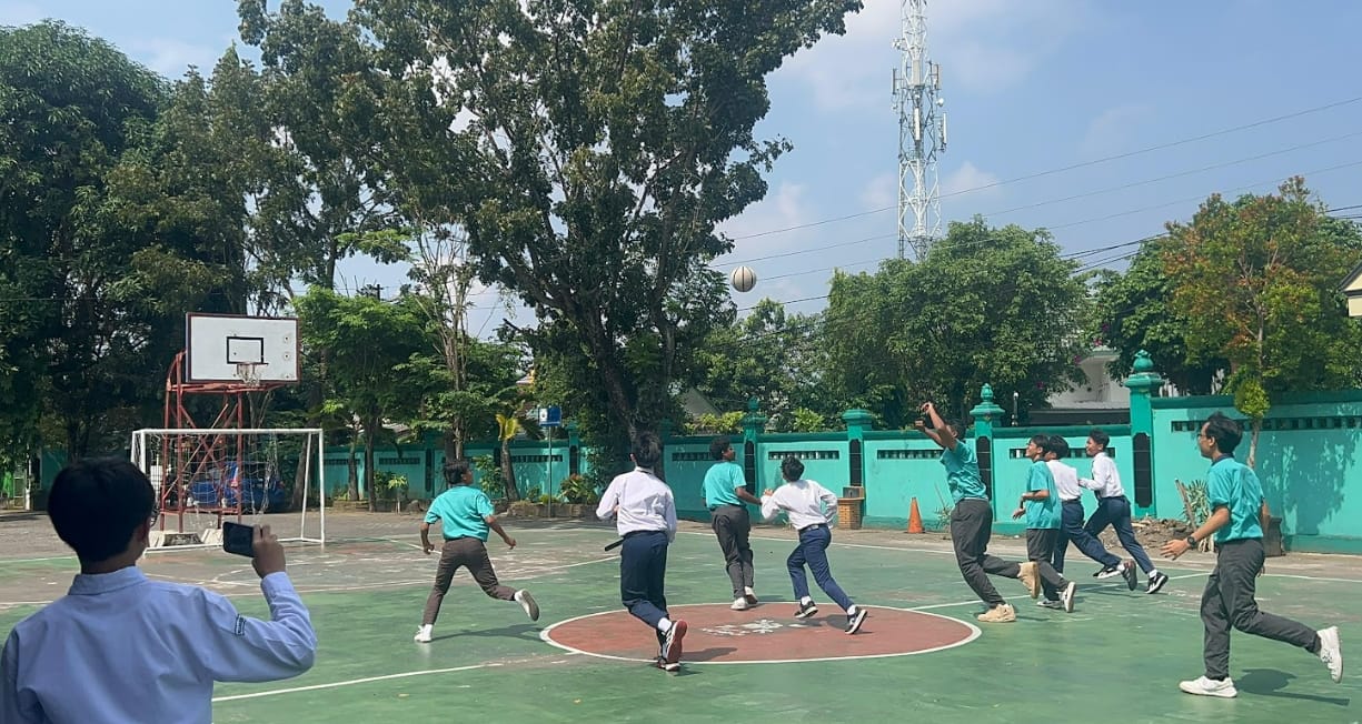 Students in teal shirts play basketball on an outdoor court while one person records with a phone.