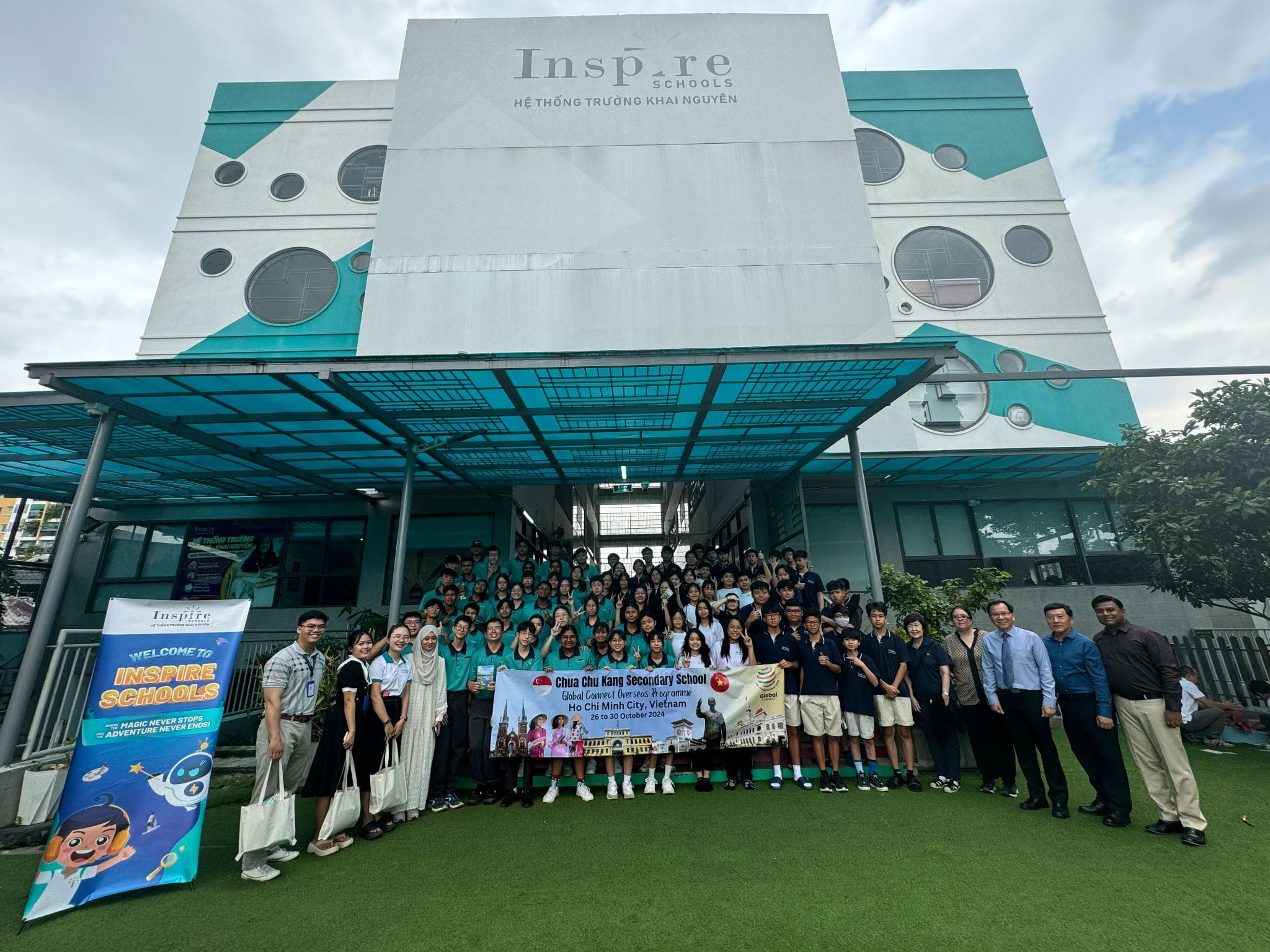 A large student group poses outside Inspire Schools in Vietnam, holding a Chua Chu Kang Secondary School banner.